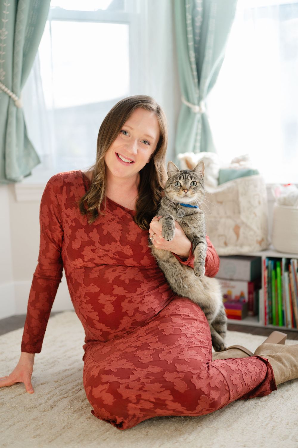 Pregnant woman in red dress sits on floor holding tabby cat in bright nursery room.