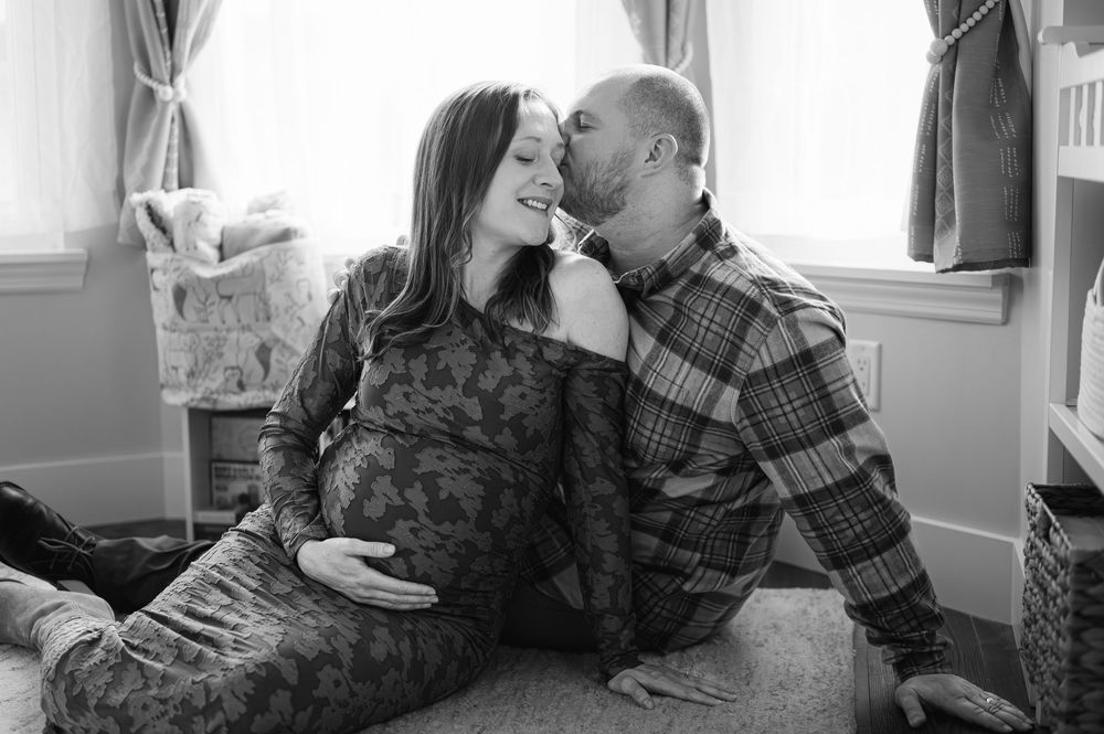 Black and white photo of pregnant couple sharing a tender kiss while sitting together in a nursery.