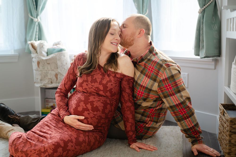 Pregnant woman in red dress sits with partner in plaid shirt, sharing a tender kiss in a cozy nursery setting.