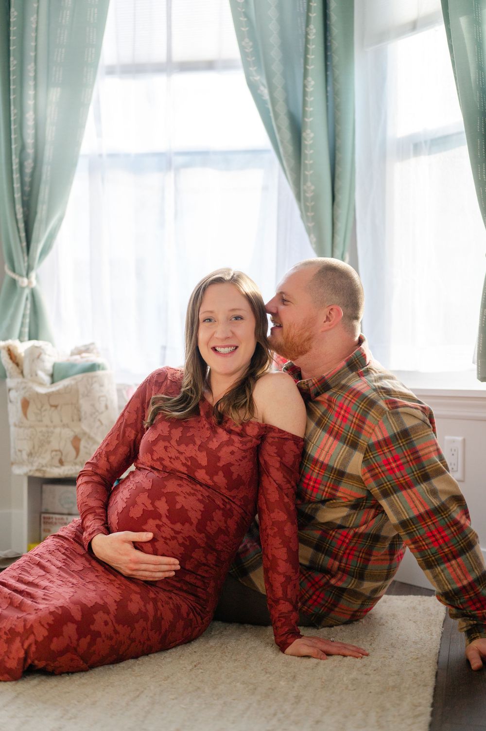 Happy pregnant couple sitting by window, man in plaid shirt whispering to smiling woman in red lace dress.