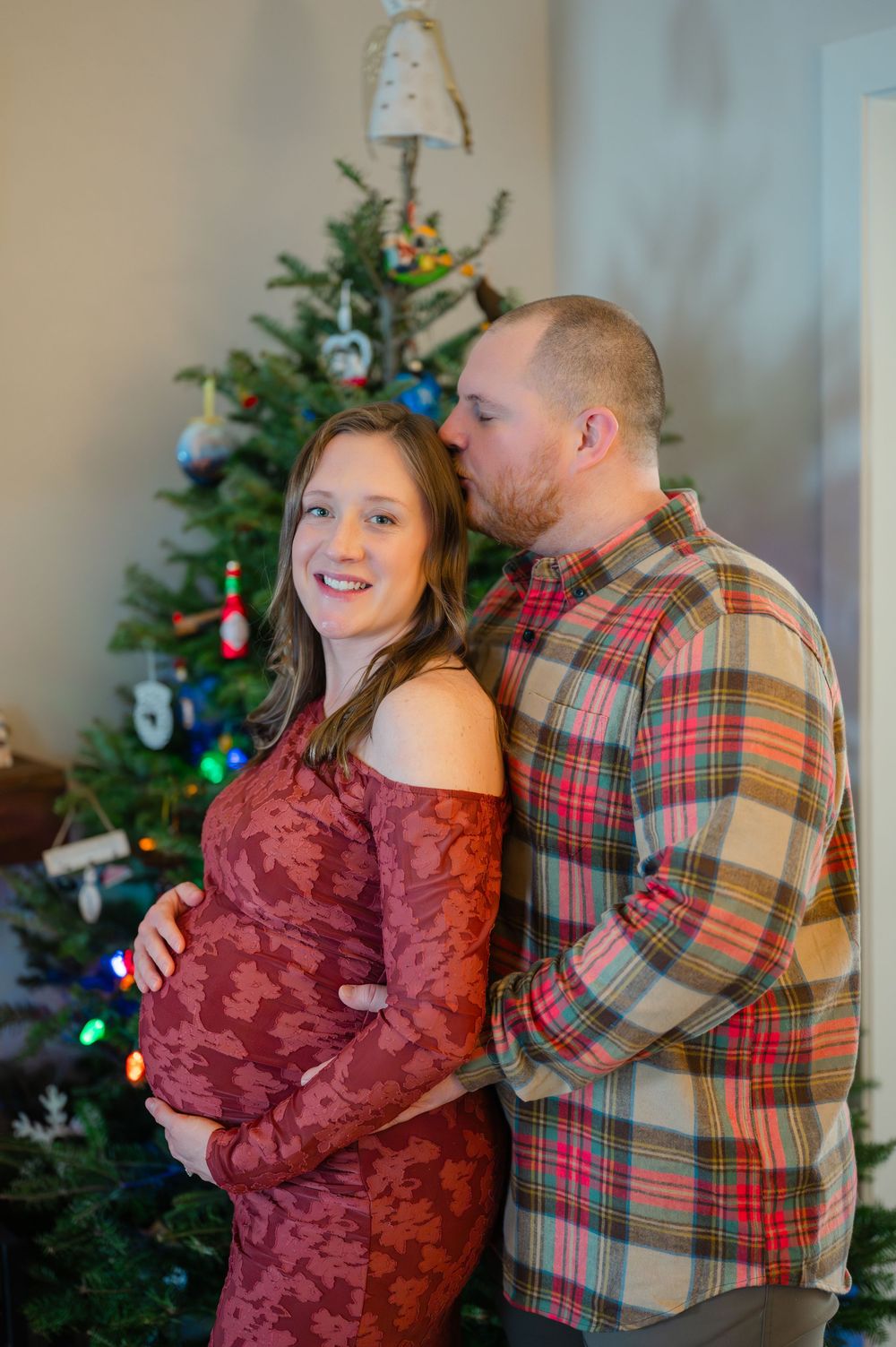 Happy pregnant couple posing in front of decorated Christmas tree, woman in red dress smiling.