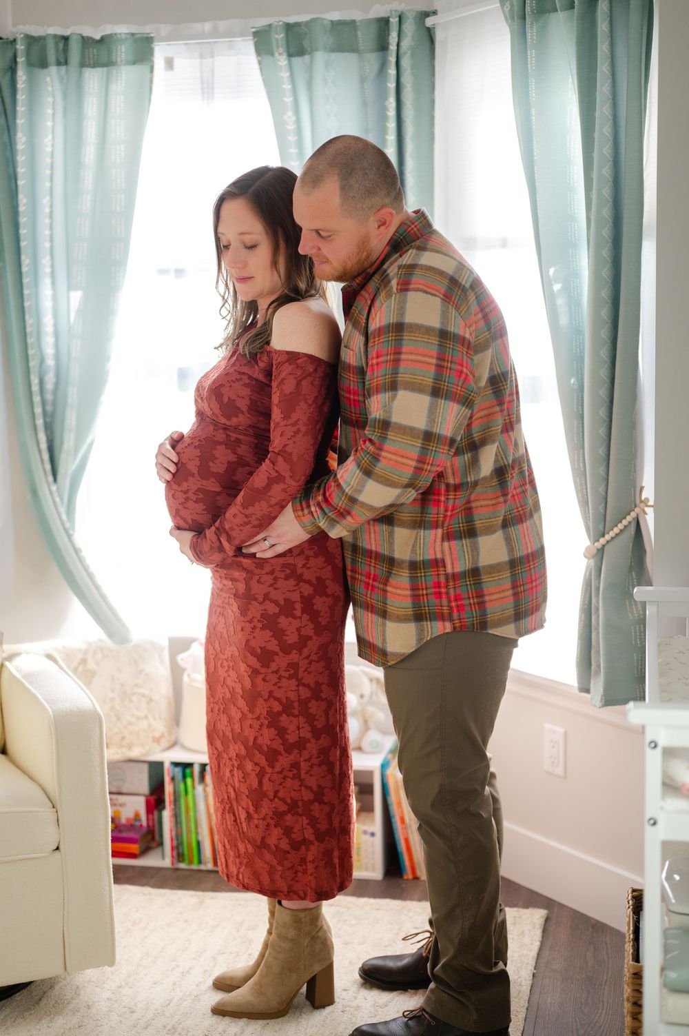 Couple shares tender maternity moment, man embraces pregnant woman in red dress by nursery window.