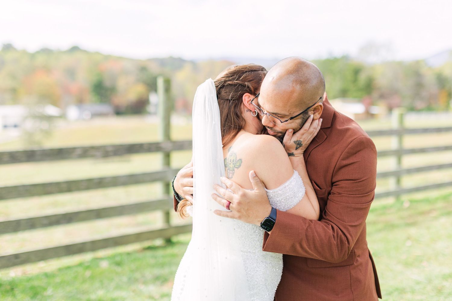 Bride and groom share a romantic first kiss outdoors near a wooden fence with fall foliage in background.