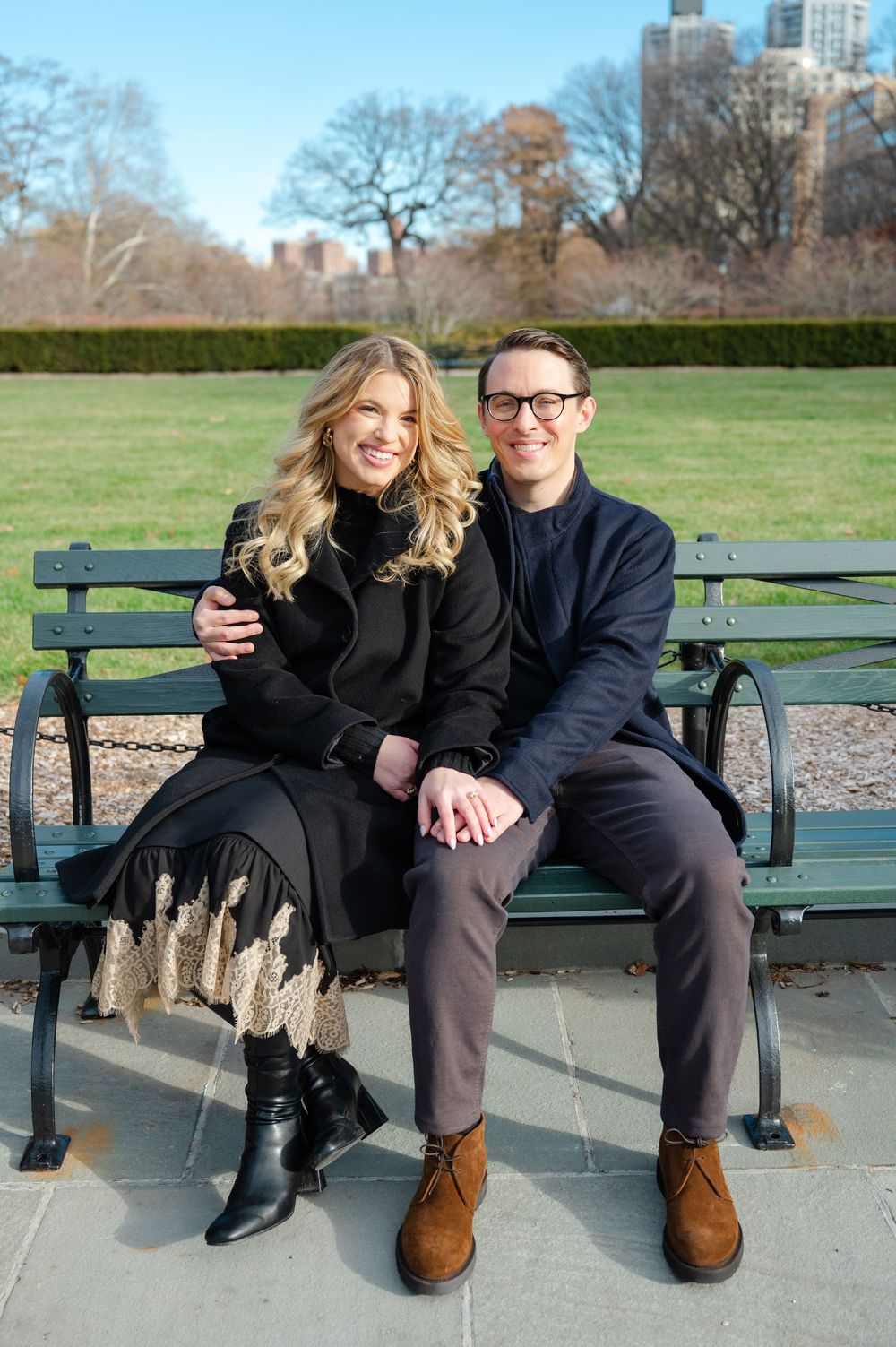 Couple smiling and sitting together on a park bench in Central Park, New York, wearing black coats on a sunny day.
