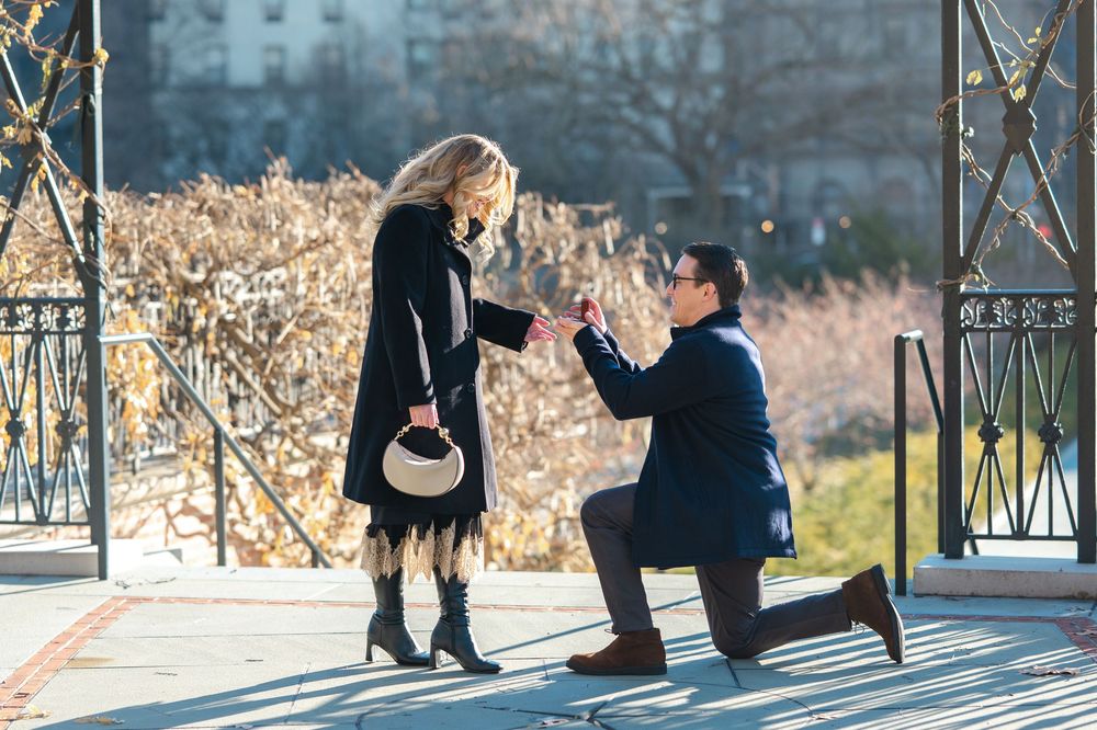 Man proposes to woman with ring in sunny park, kneeling on one knee during romantic outdoor engagement.