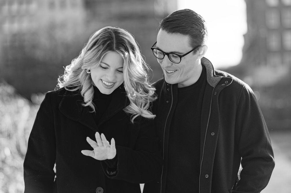 Black and white photo of a smiling couple looking at an engagement ring outdoors in winter.