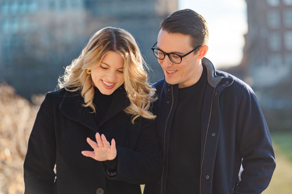 Couple in black coats admiring engagement ring outdoors in urban park setting.