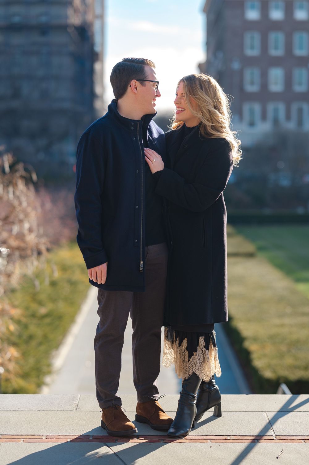 Couple in black coats sharing a romantic moment outdoors on a sunny day with brick buildings in the background.