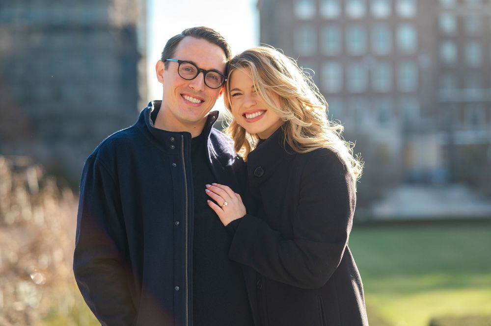 Smiling couple posing outdoors in black jackets with a city park background on a sunny day.