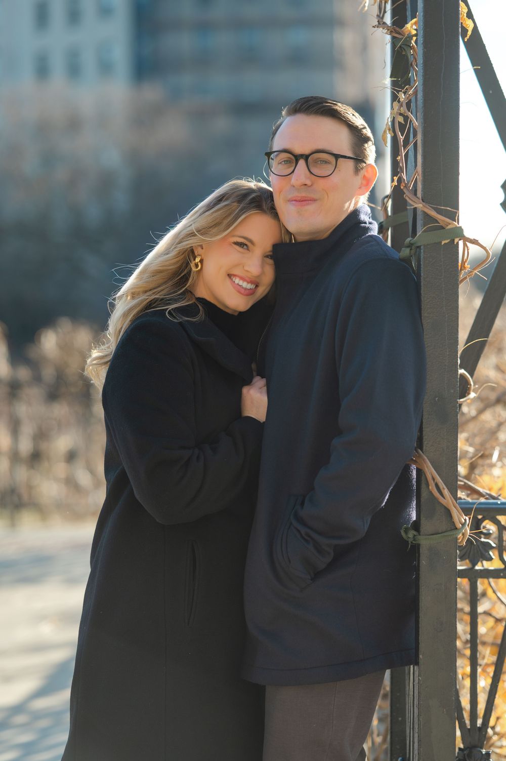 Happy engaged couple embracing by iron gate in sunny urban park, woman in black coat.
