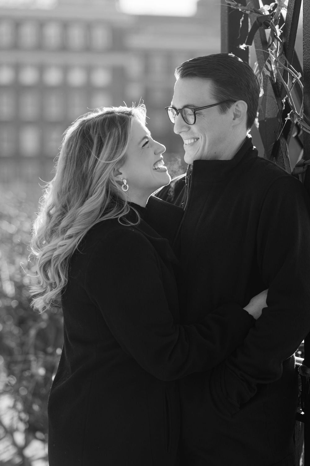 Black and white photo of a smiling couple in black coats gazing at each other outdoors.