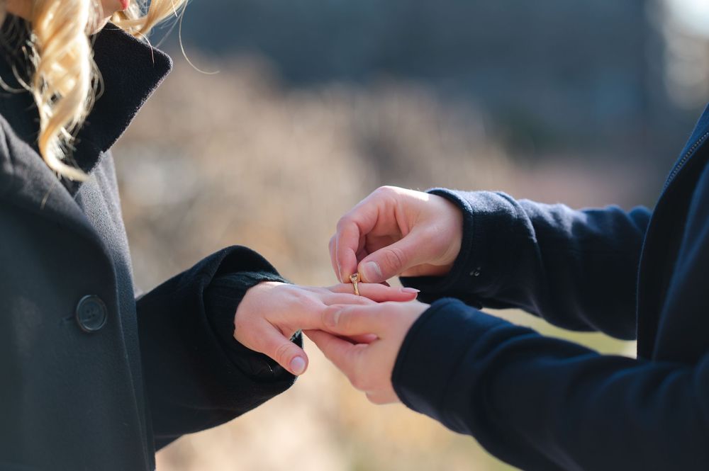 Person placing a gold ring on another person's finger outdoors during a romantic marriage proposal.