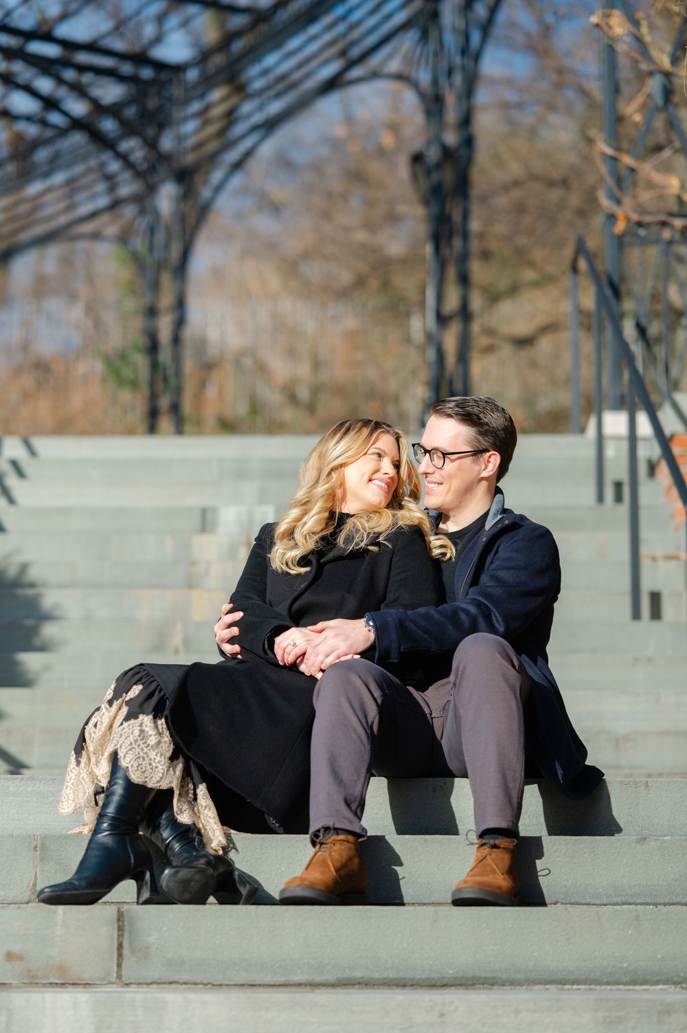 Happy couple sits on stone steps outdoors, embracing and smiling at each other during engagement photos.