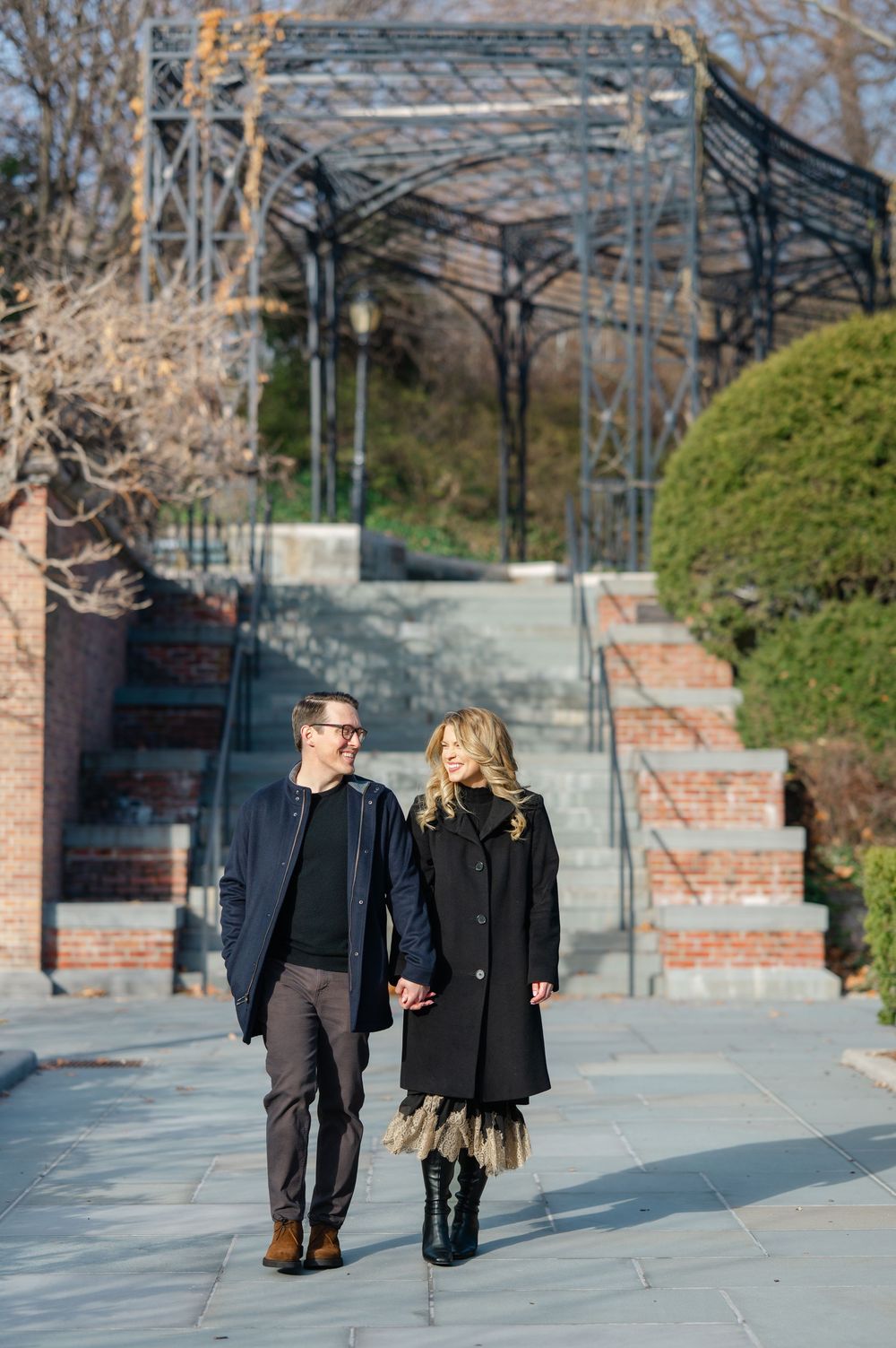 Couple holds hands walking on stone path in urban park with iron pergola structure in background.