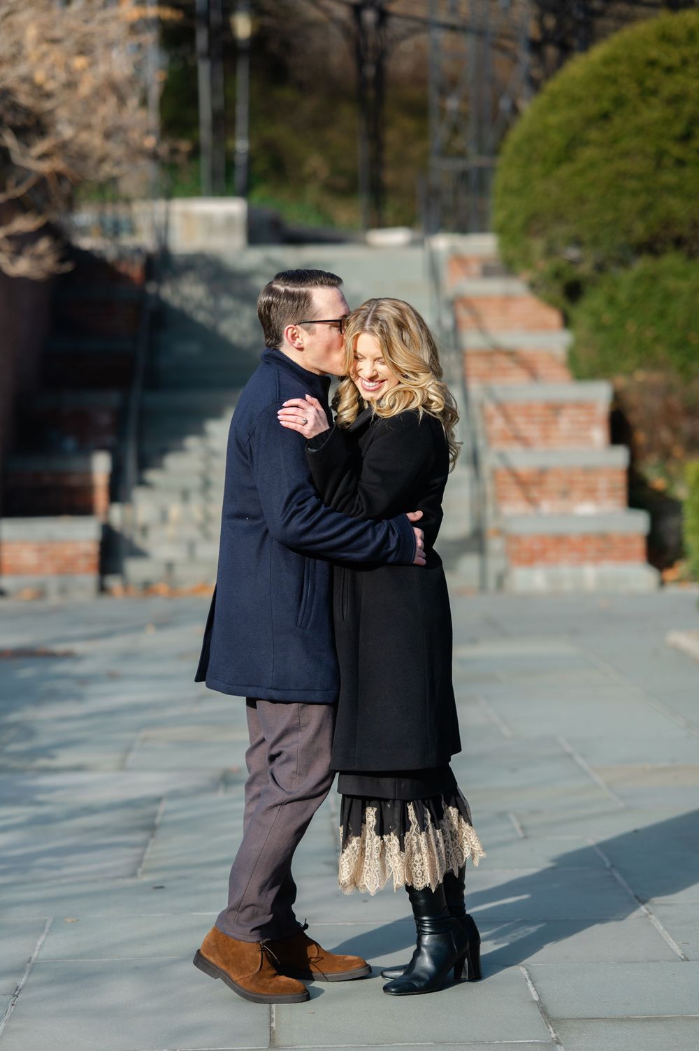 Couple embraces and kisses on stone plaza in park with brick staircase and greenery behind them.