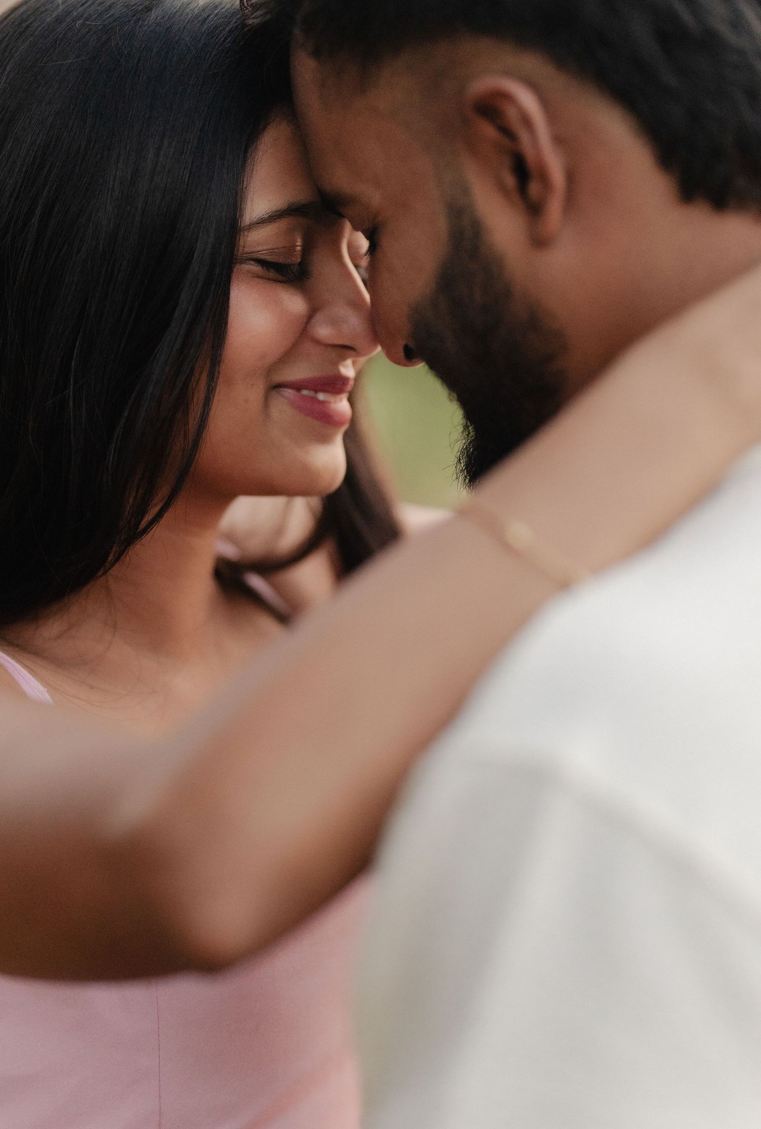 A couple shares a tender romantic moment, foreheads touching, eyes closed, in a soft outdoor setting.