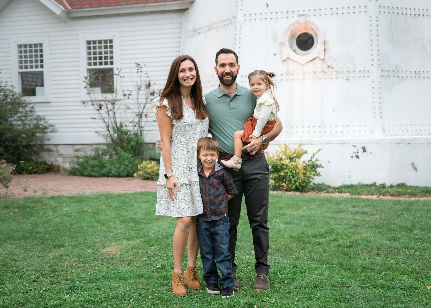 A happy family of four poses outdoors on green grass in front of a white building on a sunny day.