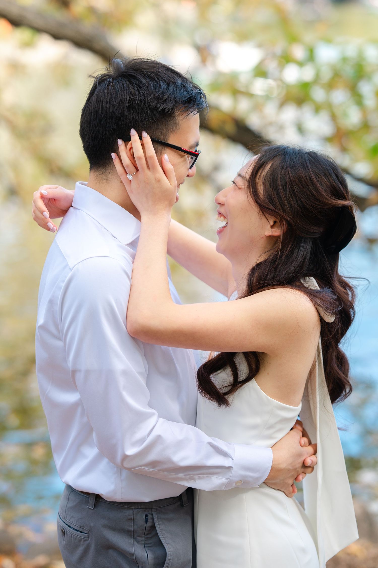 A joyful Asian couple in wedding attire embrace outdoors, the bride laughing as she touches the grooms face in Central Park.
