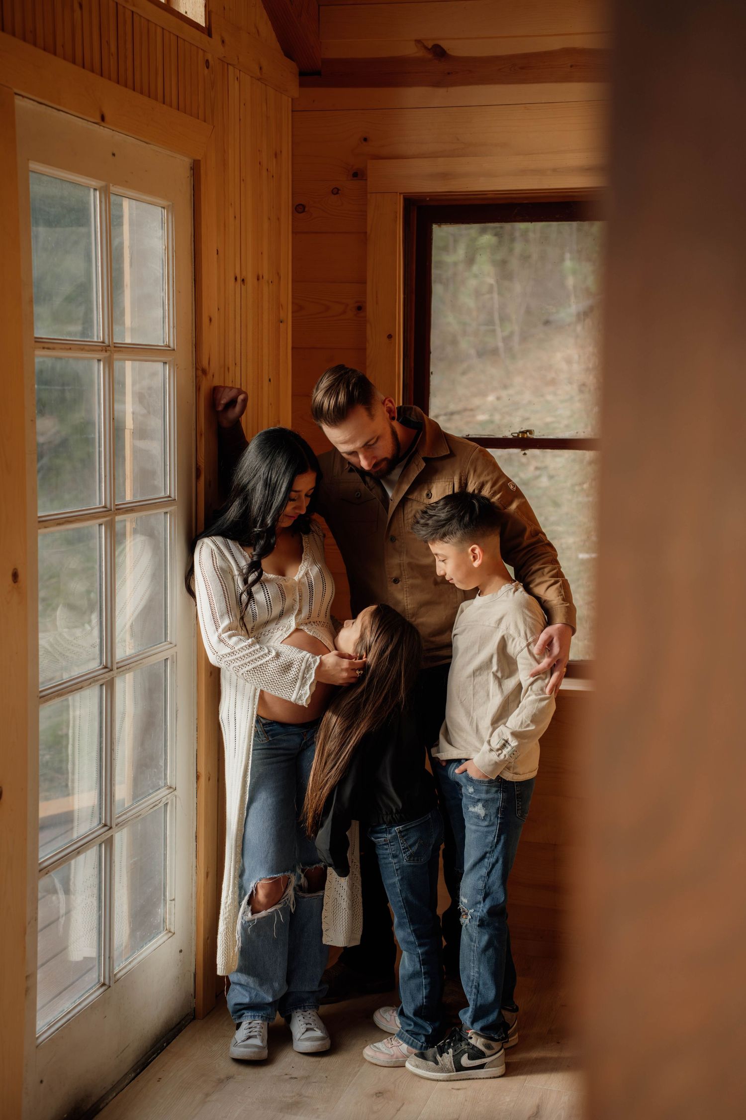 A family of four shares a warm moment in a cozy wooden cabin, gathered near a glass door with natural light.