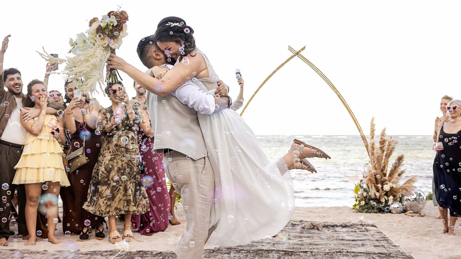 Couple sharing a romantic kiss at a beach wedding ceremony with guests celebrating around them.