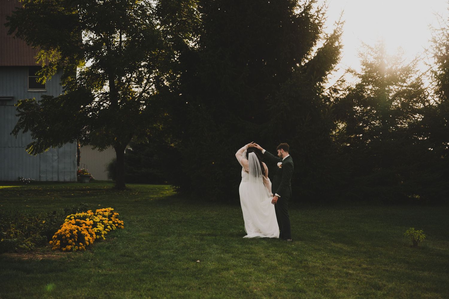 A wedding couple stands in silhouette against a sunset sky with trees and flowers in a garden setting.