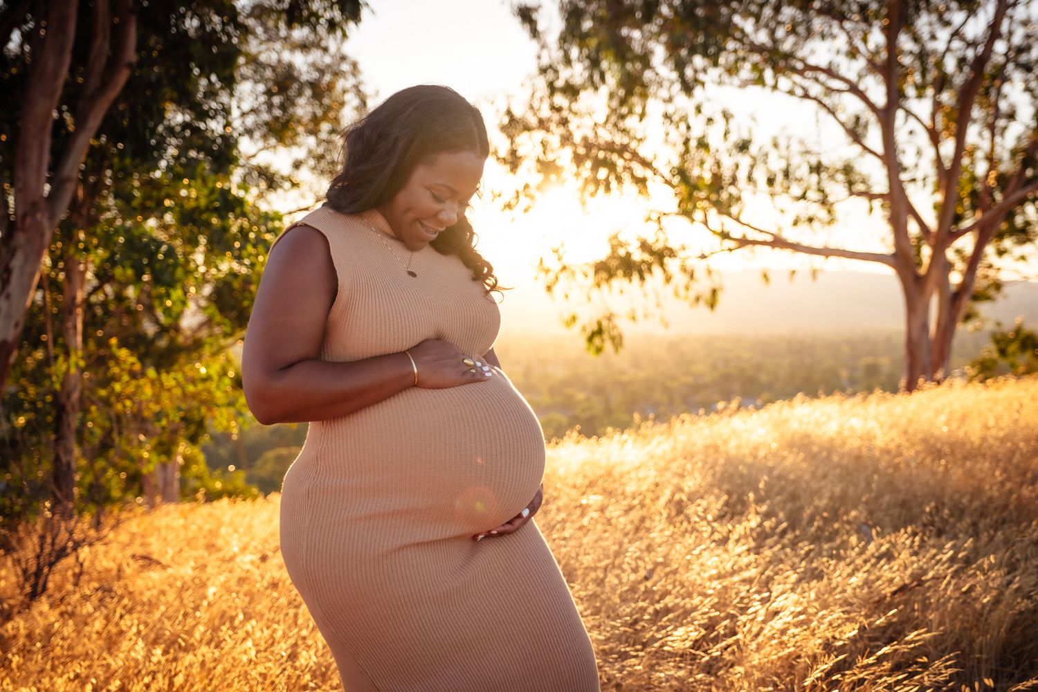 Pregnant woman in pink dress holding baby bump outdoors in golden sunlight among tall grass and trees.