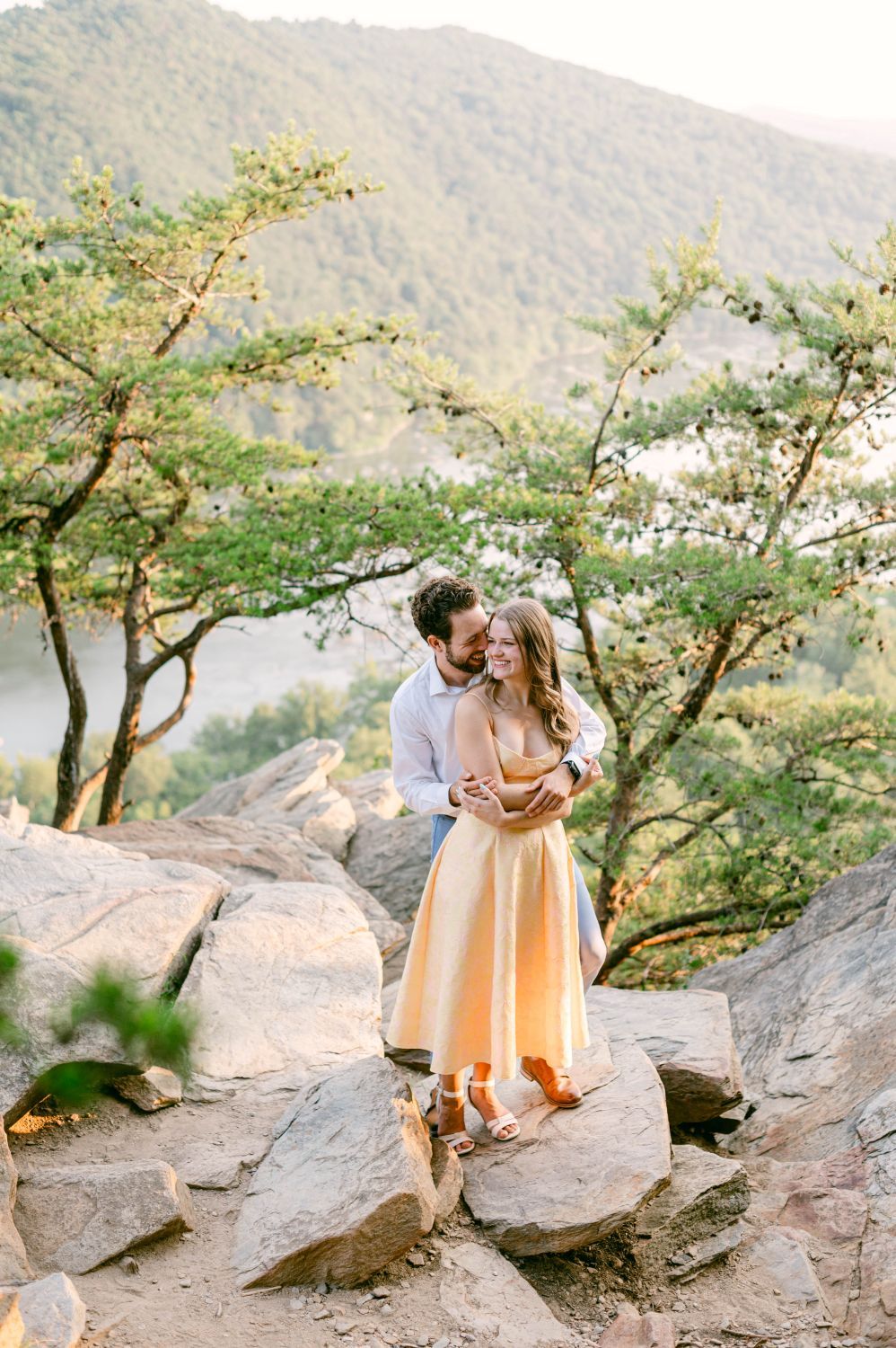Couple embracing on rocky overlook surrounded by lush green trees and mountain views during golden hour.