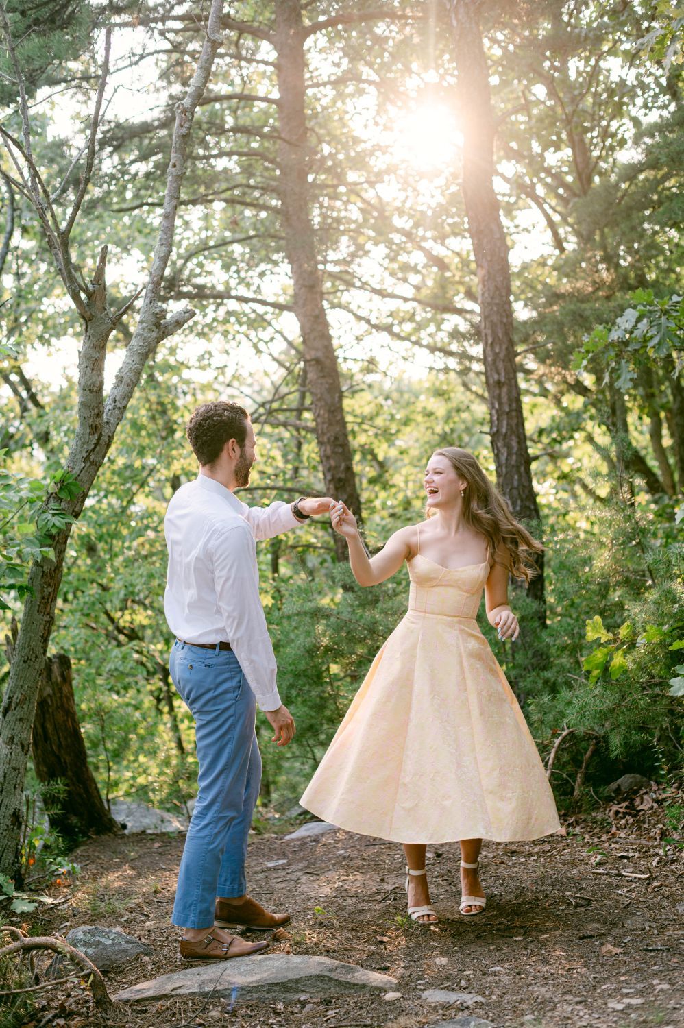 Couple dancing joyfully in sunlit forest, woman in gold tea-length dress, man in white shirt and jeans.