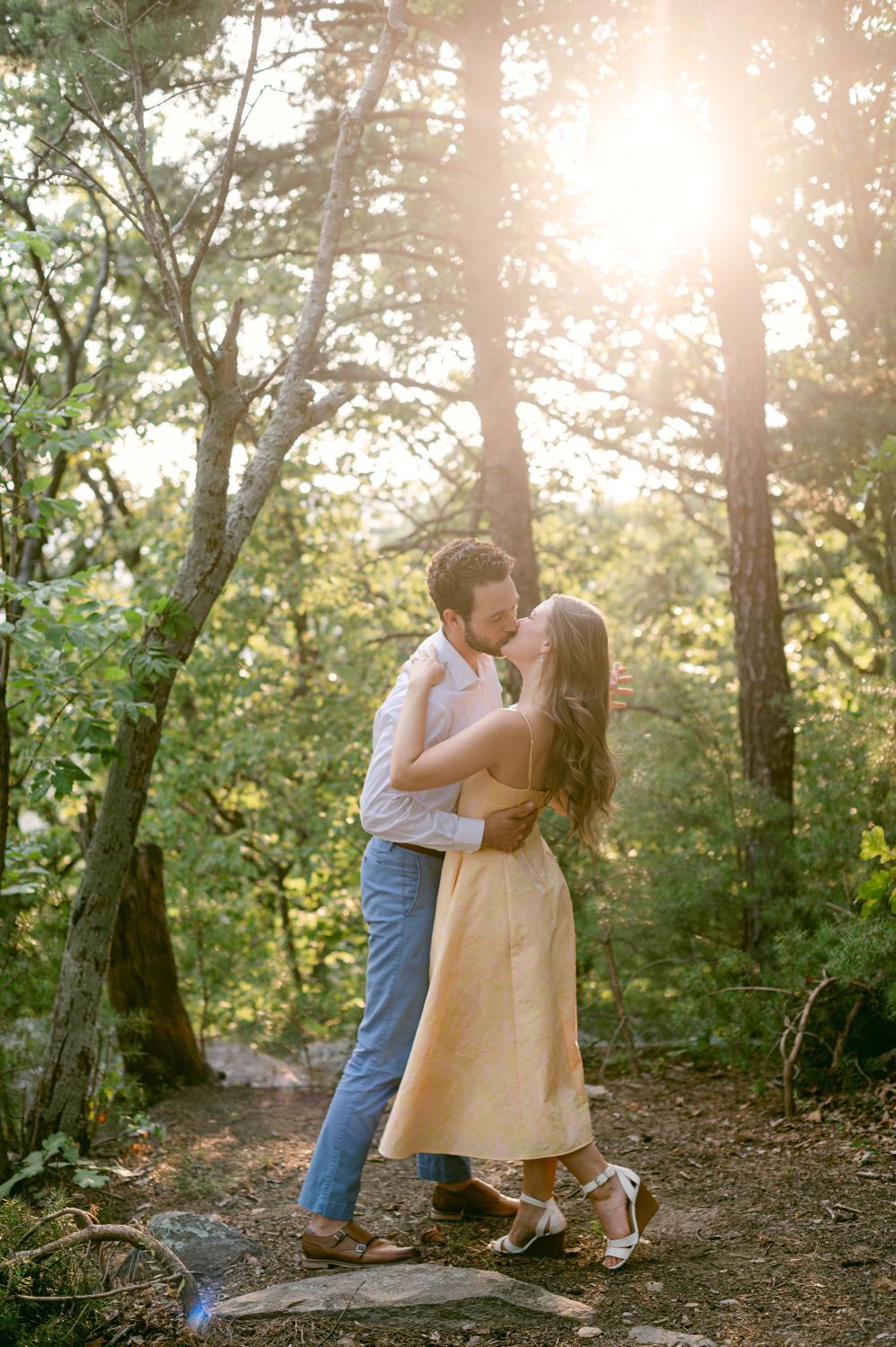 Couple sharing a romantic kiss in a sunlit forest clearing, woman in yellow dress and man in blue pants.