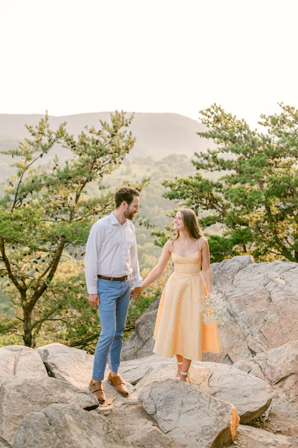 Couple holding hands on rocks with mountain views, woman in yellow dress, man in blue pants and white shirt.