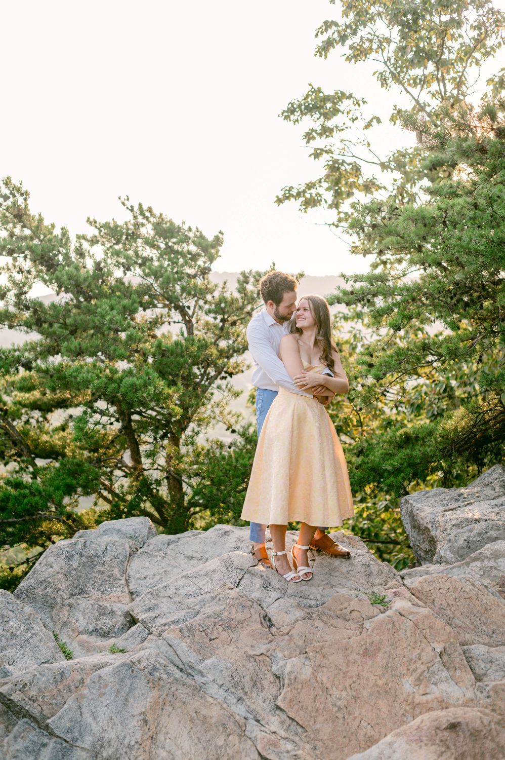 Couple embracing on rocky overlook surrounded by lush green trees during golden hour engagement session.