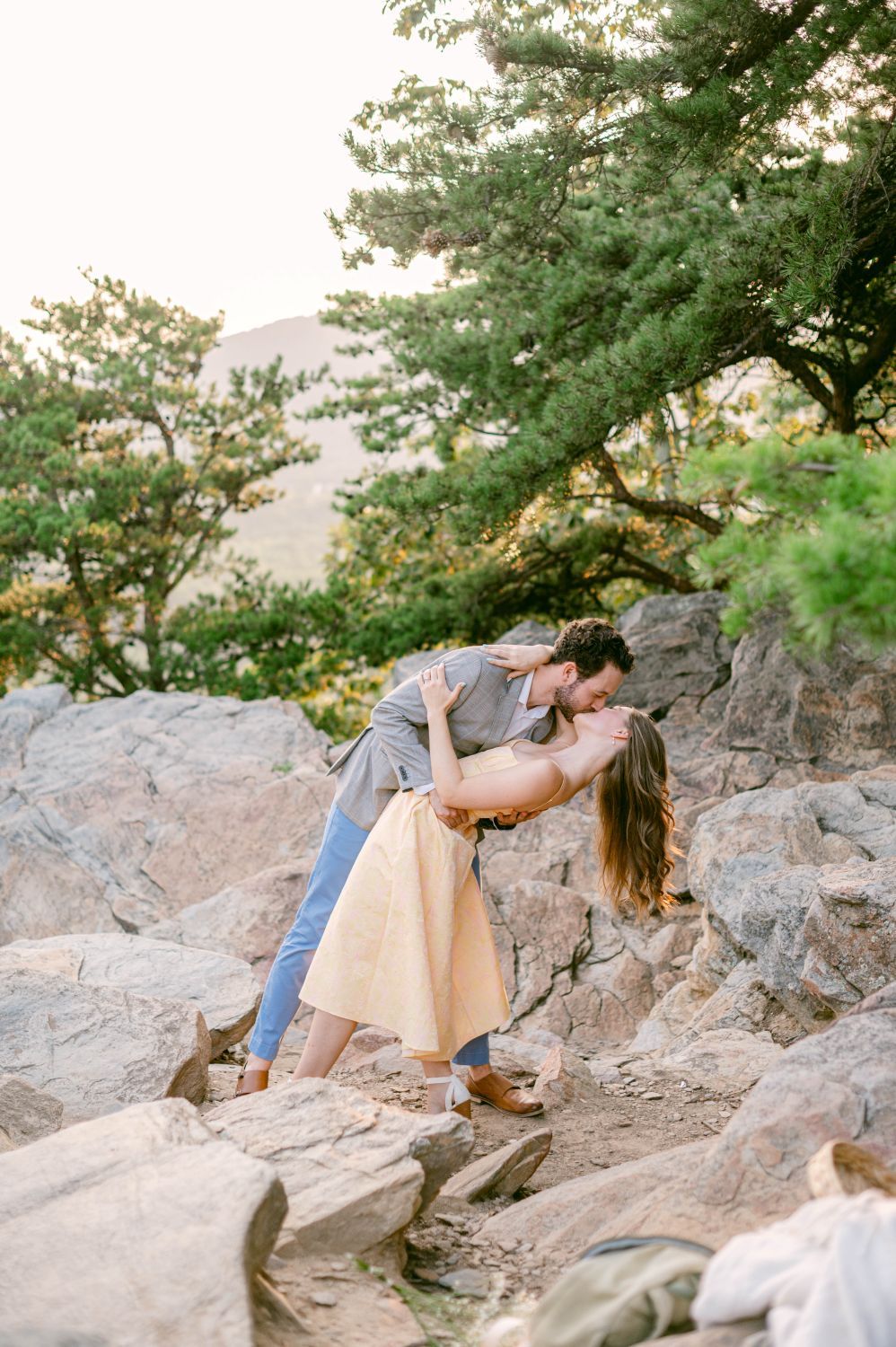 Couple sharing a romantic dip kiss on rocky terrain surrounded by lush green trees during an outdoor engagement session.