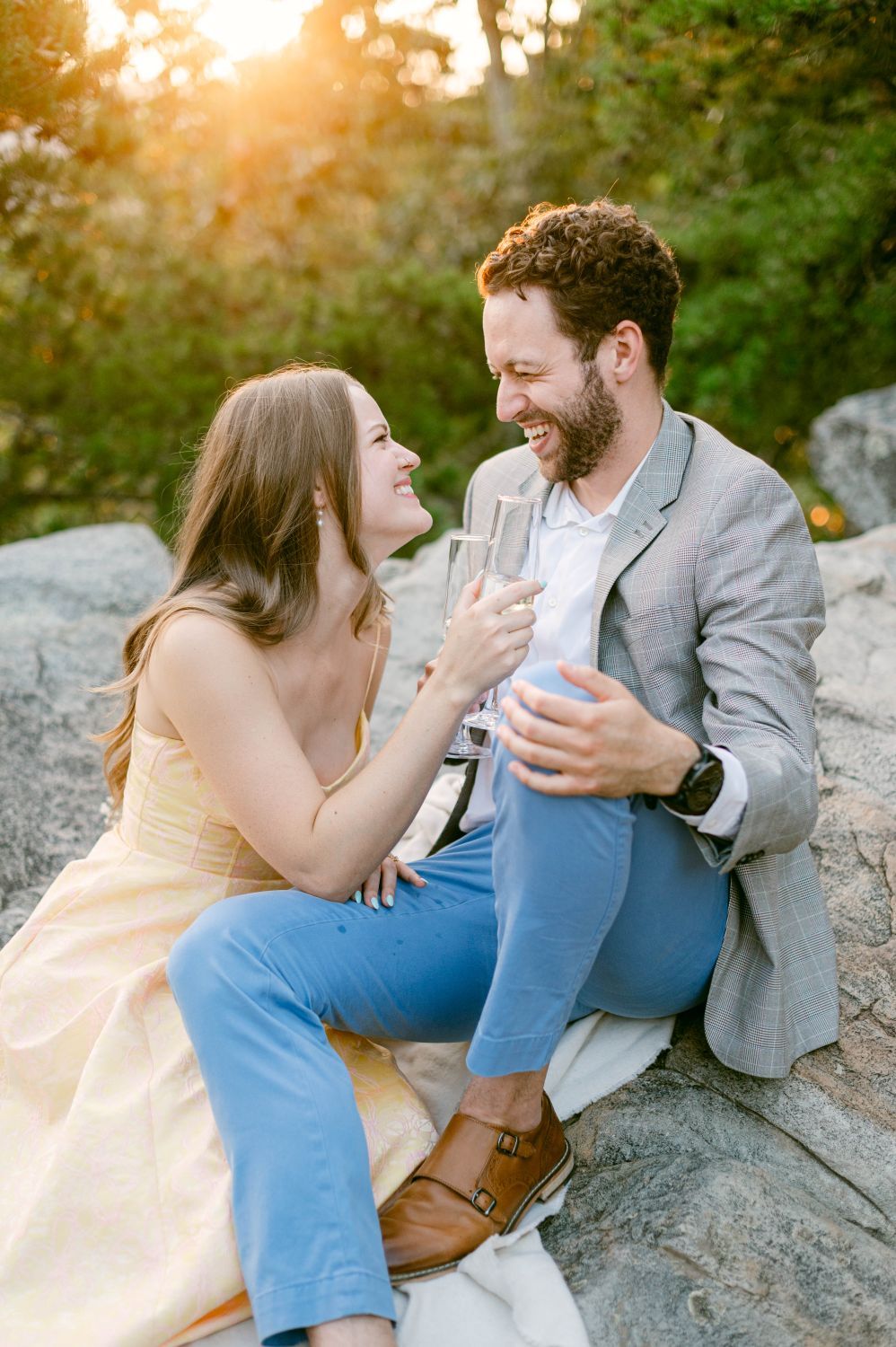 Couple laughing and toasting with champagne glasses while sitting on rocks during golden hour outdoors.