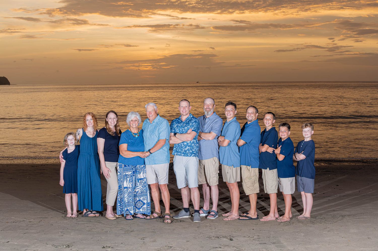 Eight people holding hands on a beach at sunset, silhouetted against a vibrant orange and blue sky over calm waters in Tamarindo