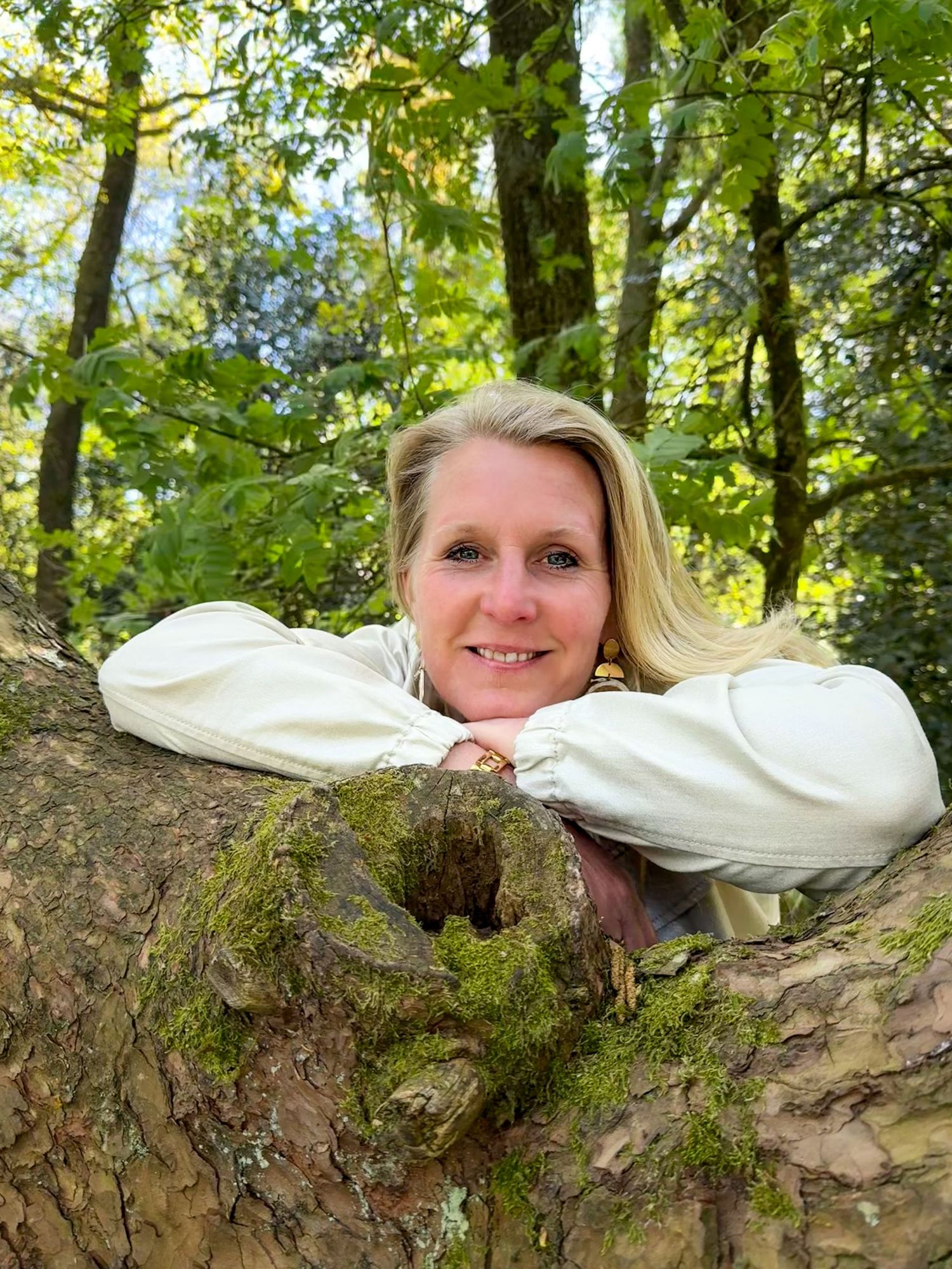 Smiling blonde woman resting on moss-covered tree trunk in green forest.