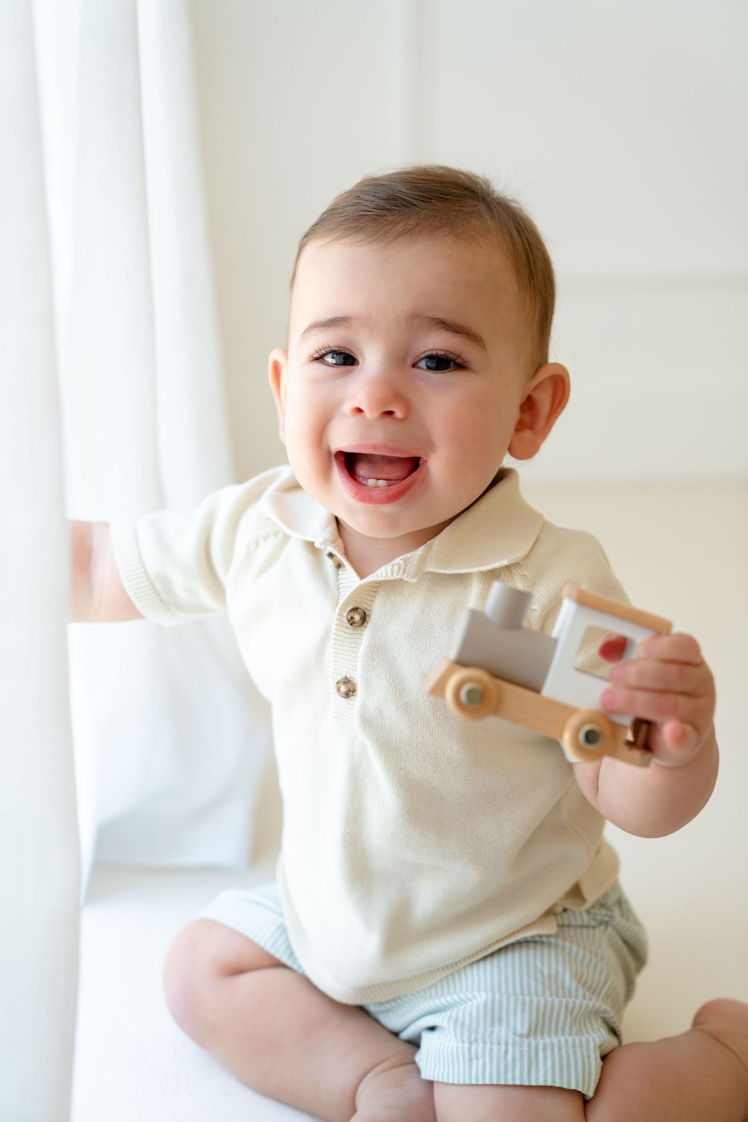 Smiling baby boy in cream romper holding a wooden toy train while sitting near a bright white window.