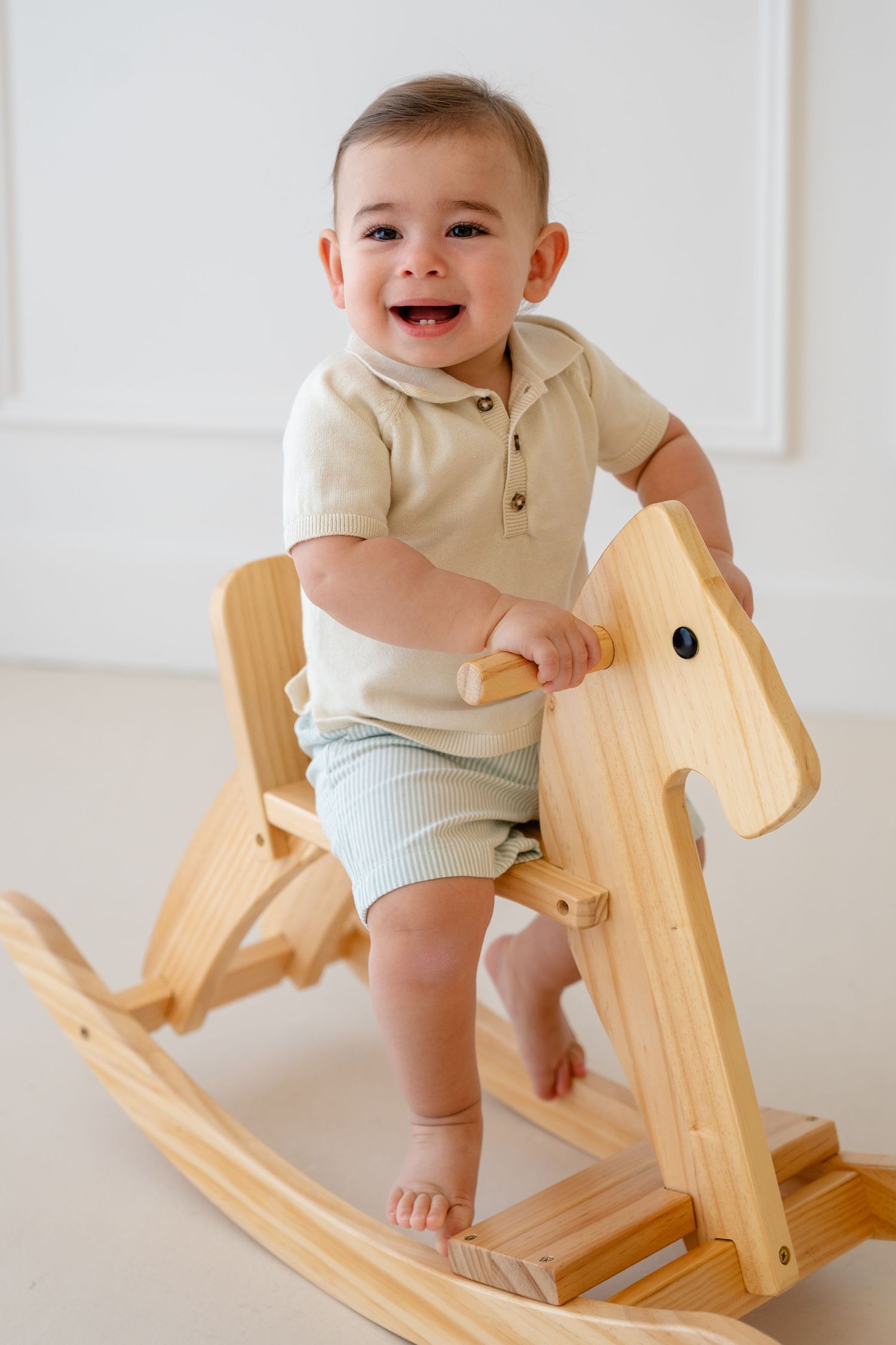 Smiling baby in beige romper standing on wooden rocking horse toy in bright white room.