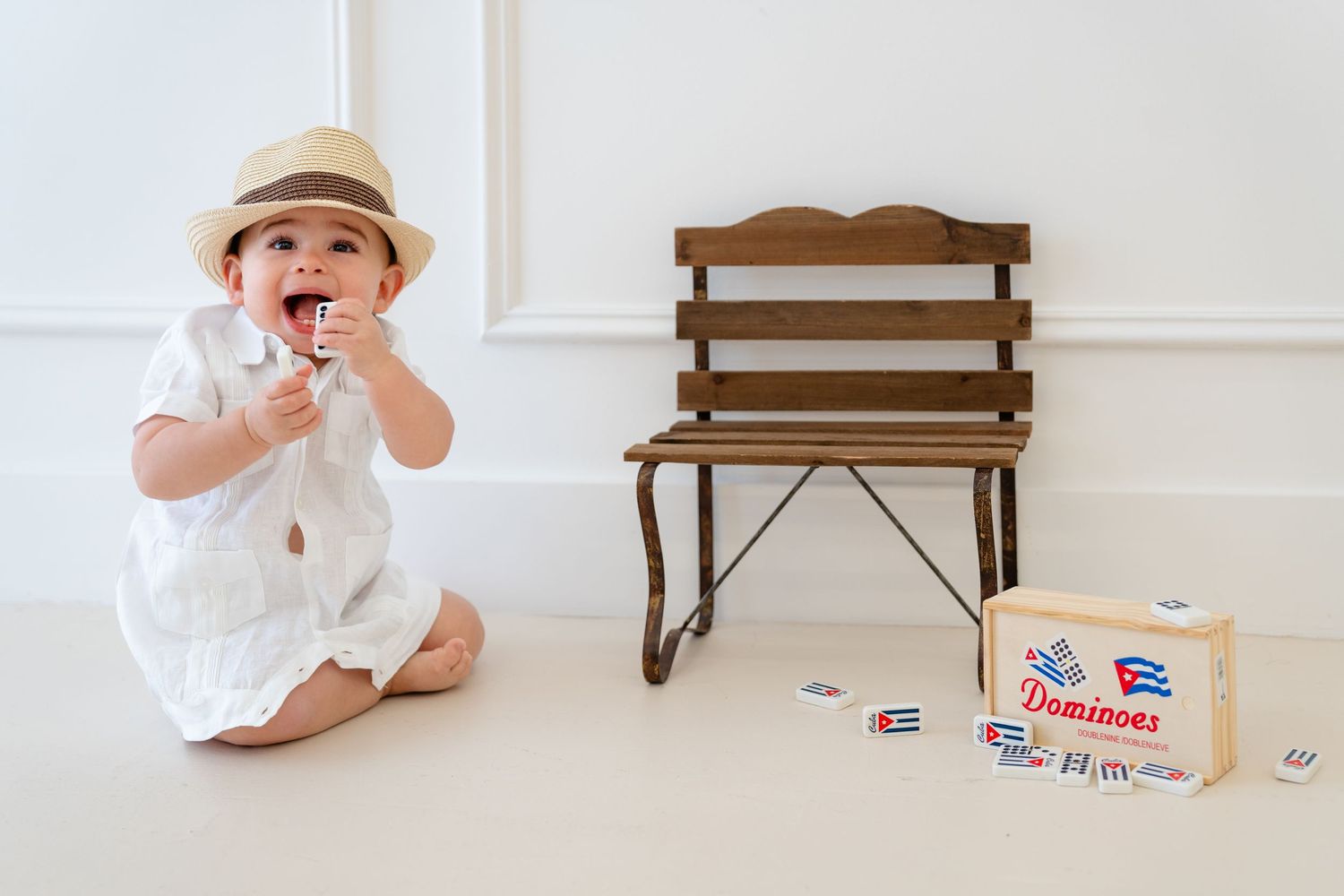 Adorable baby in white outfit and straw hat sitting on floor playing with Dominoes toy blocks.