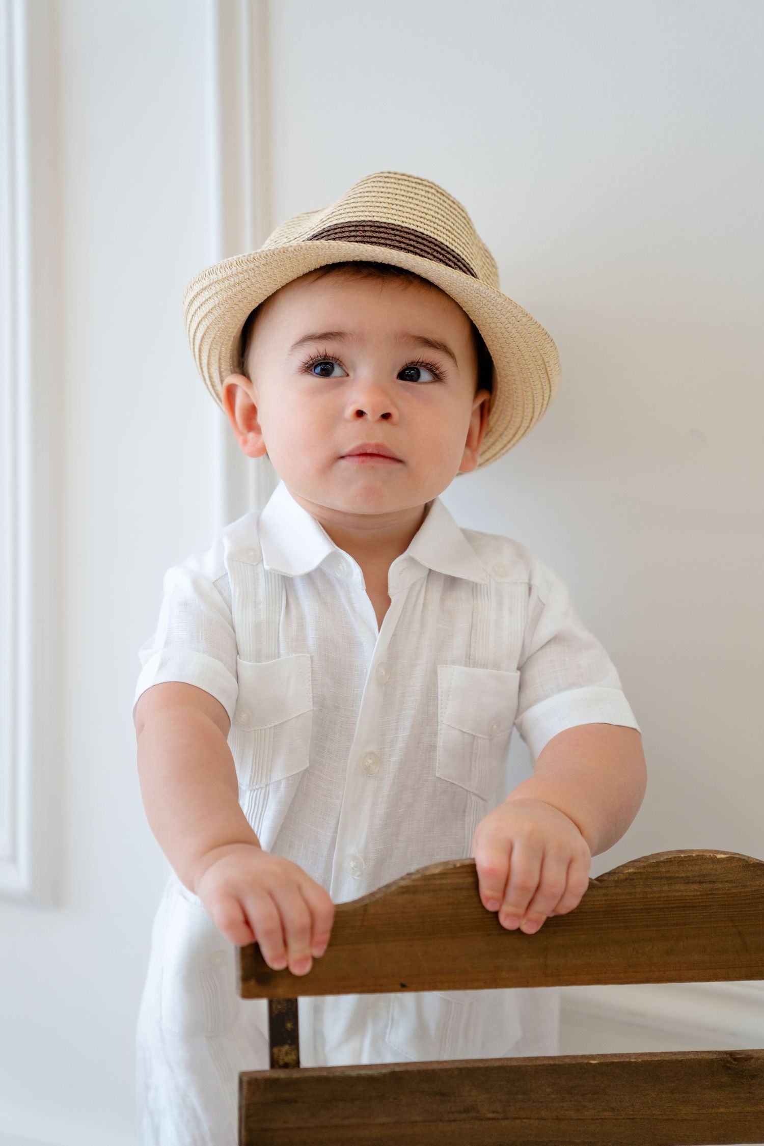 Adorable baby boy in white linen outfit and straw fedora hat standing at wooden bench against white wall.