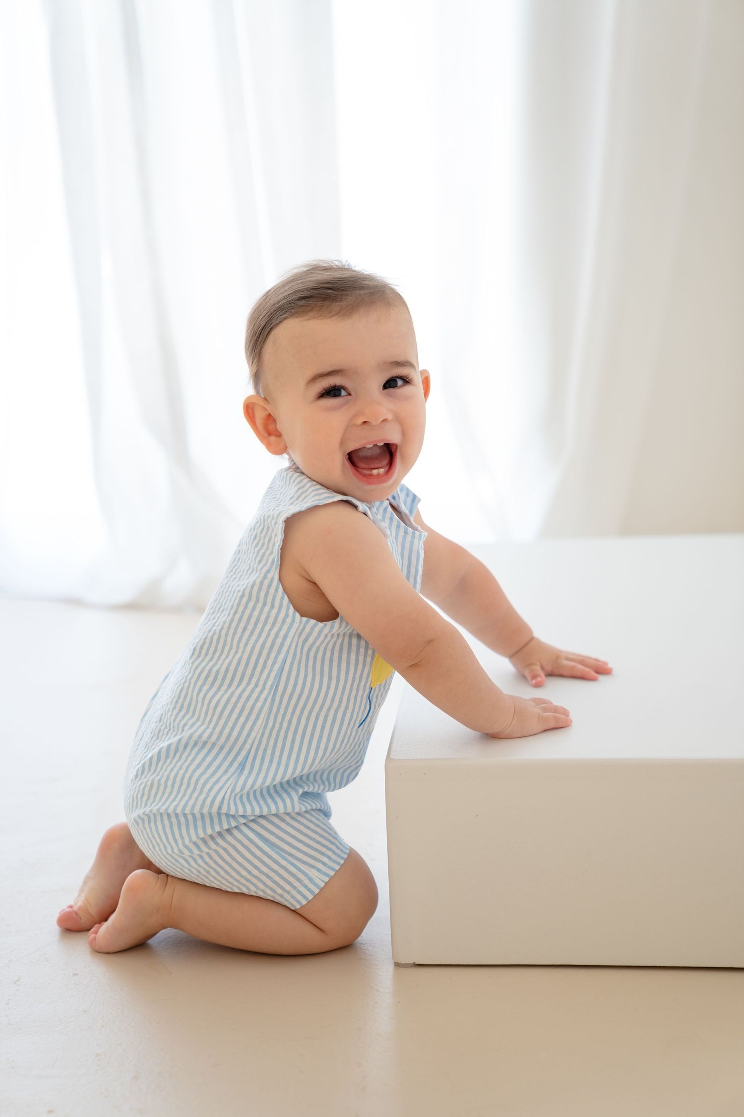 Happy baby in blue striped romper kneeling on floor, leaning on white box, smiling joyfully in bright white room.