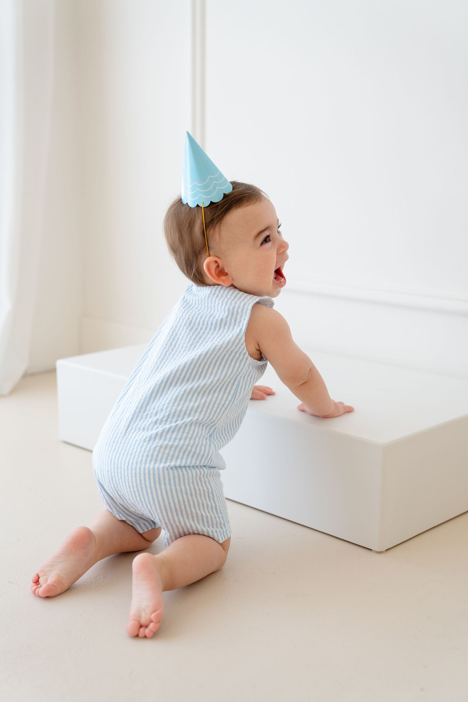 Cute baby in blue striped romper and party hat crawling near a white block in a bright room.