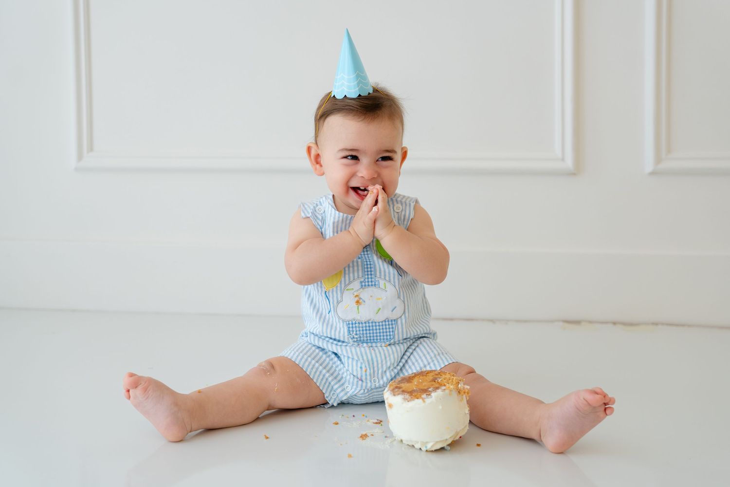 Baby in blue romper and party hat smashes first birthday cake, sitting on white floor with frosting on hands.