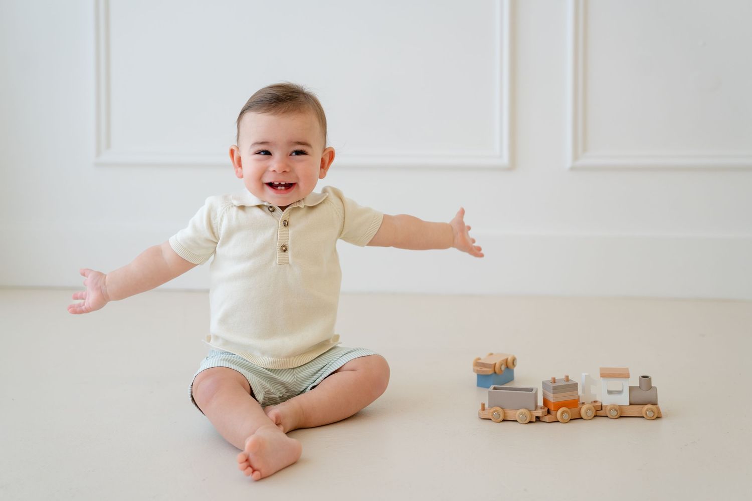 Happy baby boy in cream romper sits on white floor with arms outstretched, smiling beside a wooden toy train.