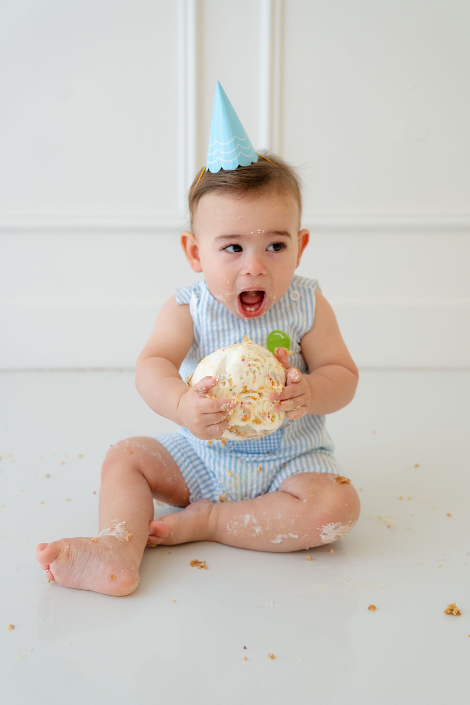 Baby in blue romper and party hat smashes first birthday cake while sitting on white floor.