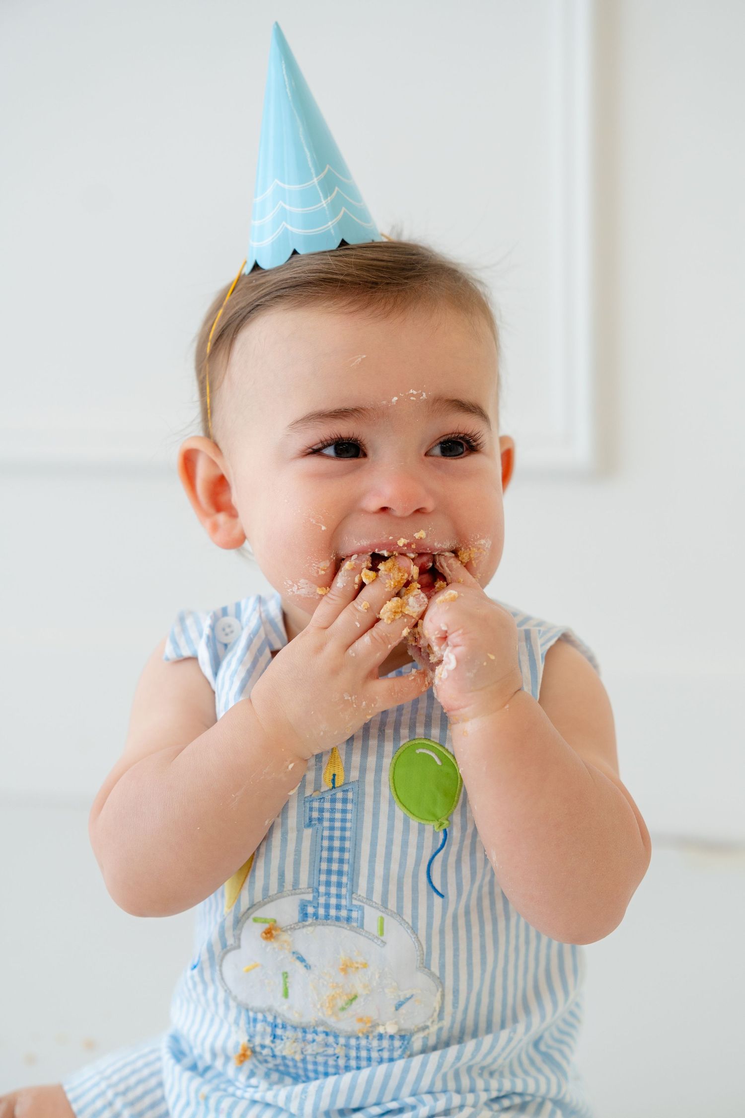 A baby in a blue party hat joyfully smashes and eats birthday cake with both hands, wearing a striped outfit.