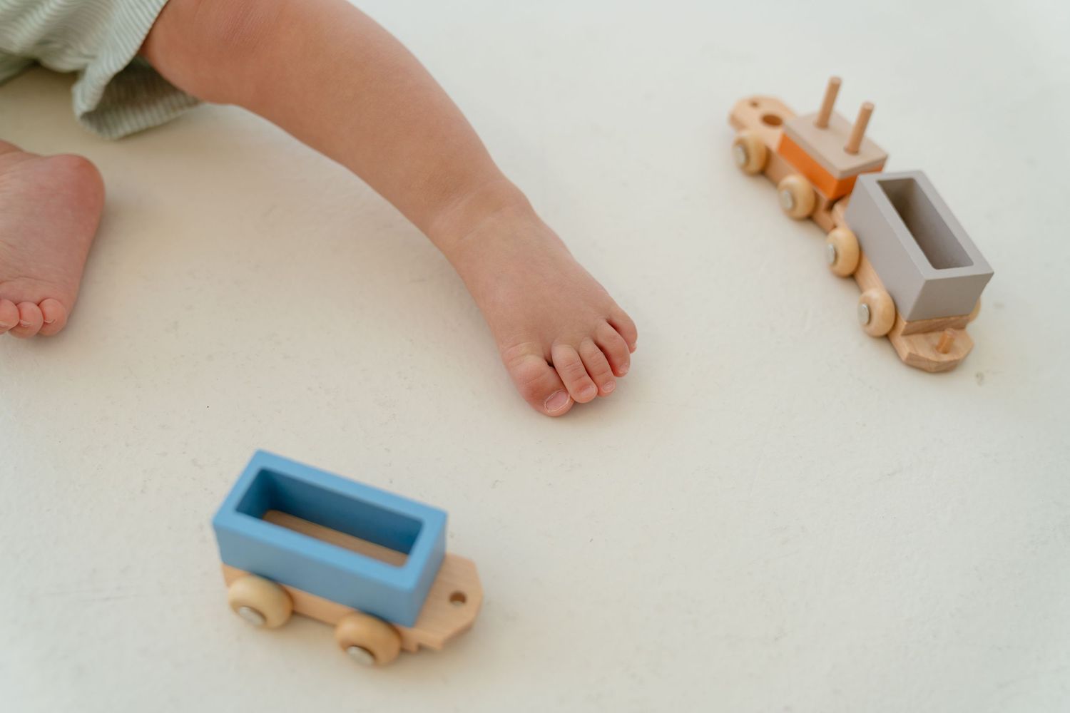 Baby's foot beside colorful wooden toy train set on white surface, perfect for toddler imaginative play.