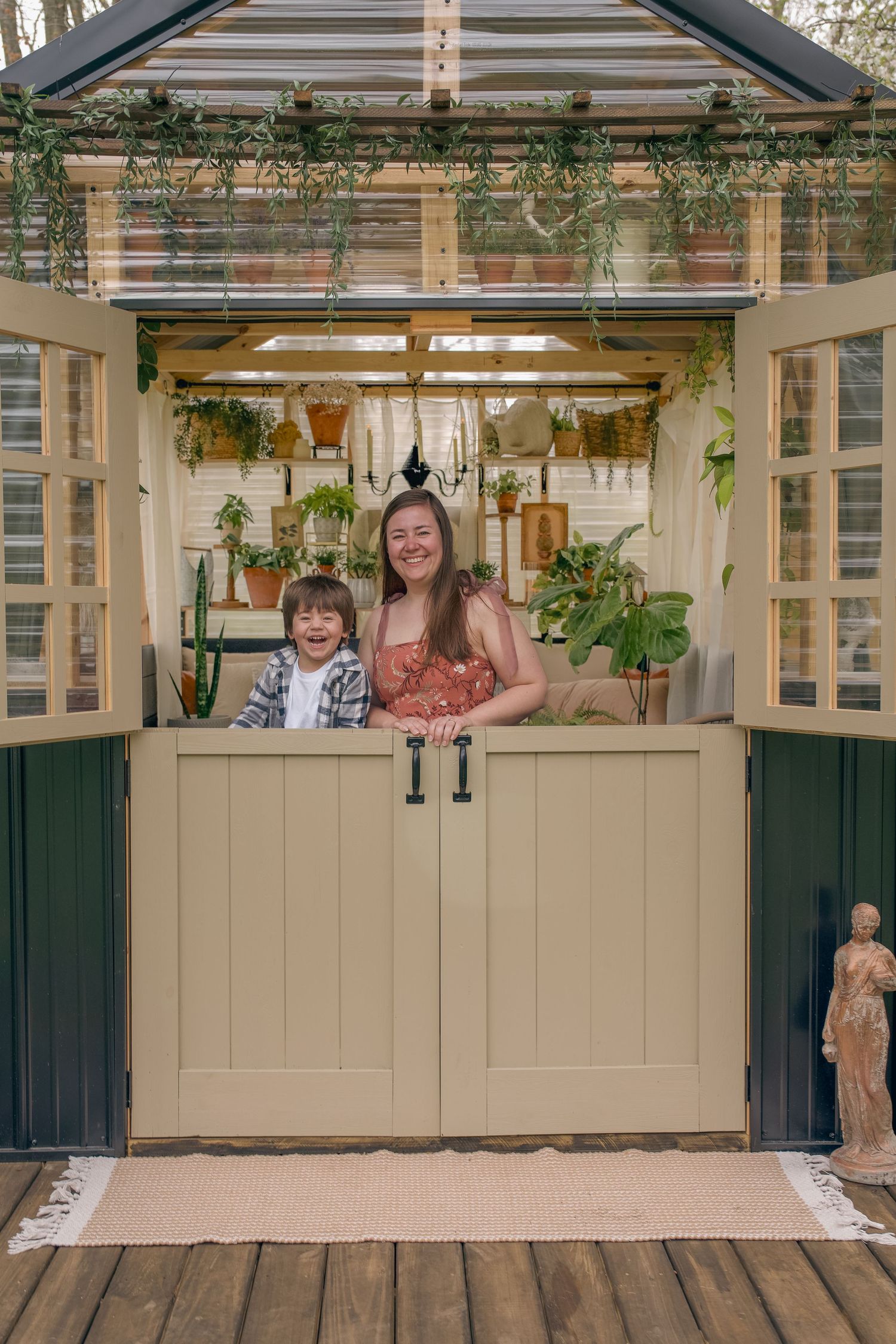 A smiling woman and young boy stand behind Dutch doors of a charming greenhouse shed filled with hanging plants.