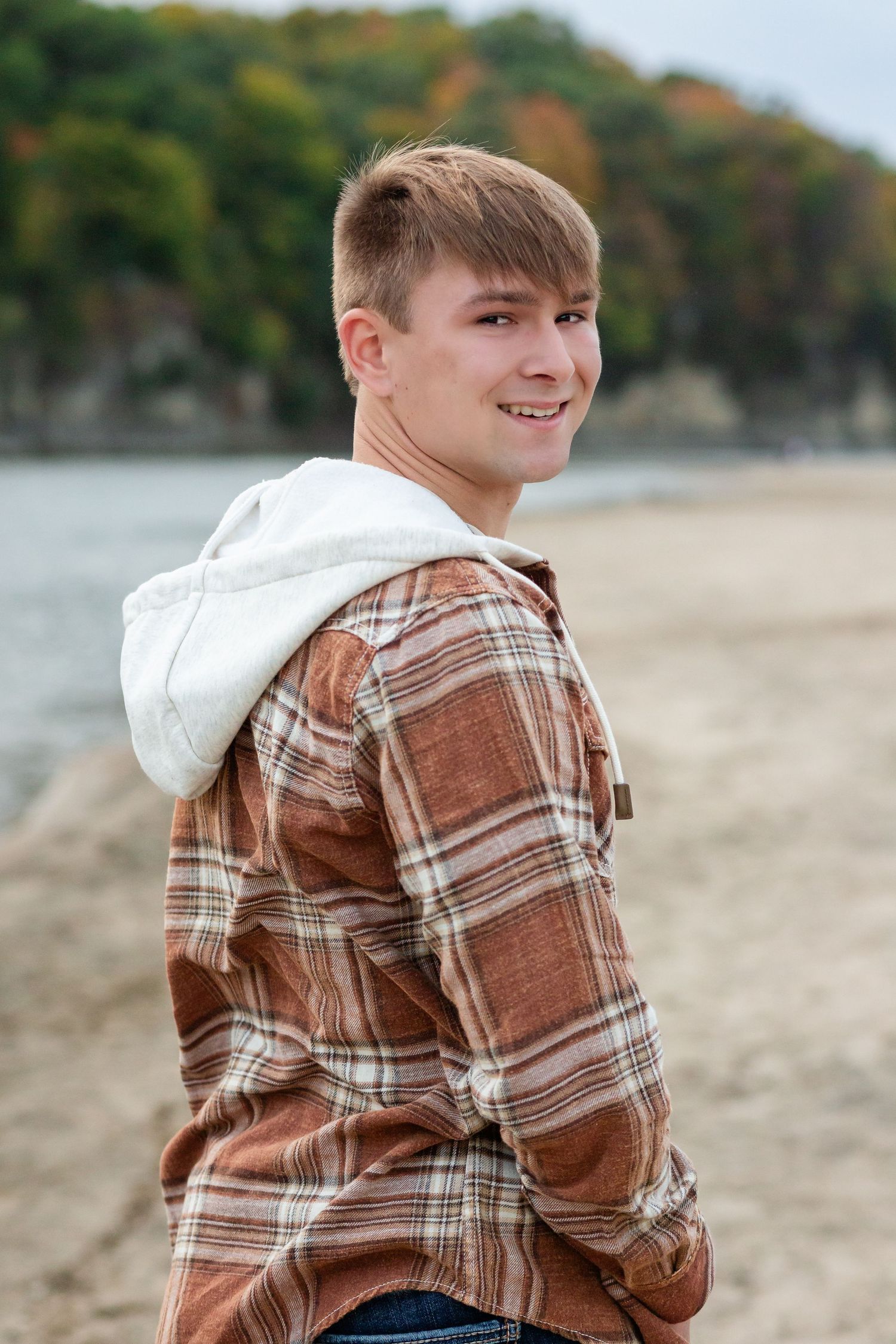 Smiling teenage boy in plaid hoodie shirt standing on a sandy beach with autumn trees in the background.