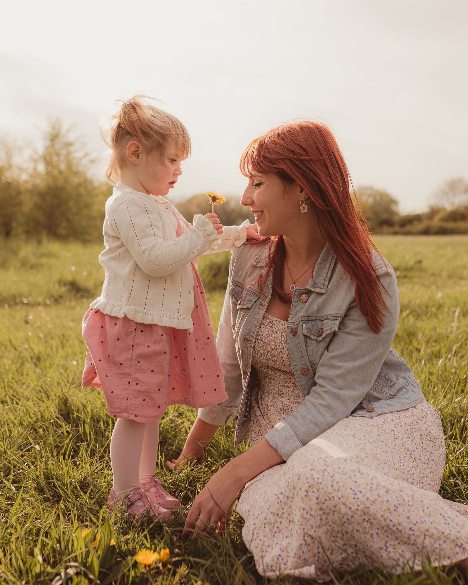A red-haired woman kneels in a sunny field as a toddler girl in a pink skirt offers her a yellow dandelion.