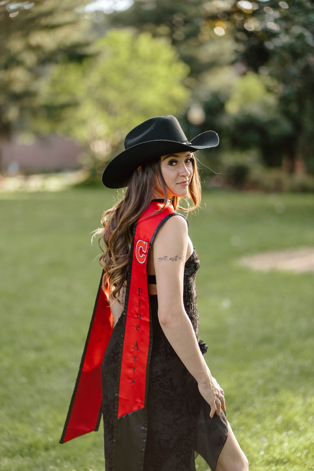 Woman in black cowboy hat and red graduation sash poses outdoors in a park setting.
